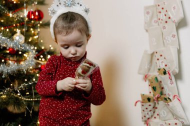 Kar tanelerinden yapılmış beyaz bir tacı olan küçük beyaz bir kız elinde çikolatalı Noel Baba ile Noel Baba paketini açarak Noel ağacı ve takvim arasında duruyor.