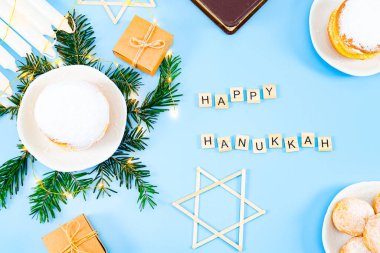 Üzerinde yazı olan tahta küpler: Mutlu Hanukalar, çörekler, İncil, mumlar, köknar dalları ve açık mavi arka planda bir çelenk..