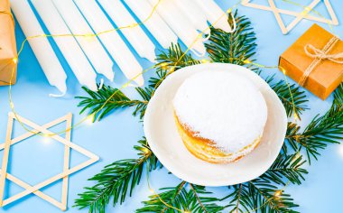 One large donuts with powder and cream in a saucer on fir branches, nine candles, a garland, stars of David and craft boxes of gifts lie on a blue background, flat lay close-up. Hanukkah Celebration clipart