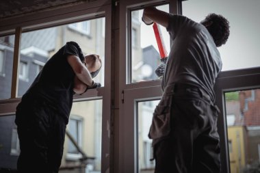 İki yakışıklı beyaz adam iskelede yarı yarıya dönerek pencereyi elle onarıyor. Plastik çerçevelerdeki çatlakları yay üzerinde silikon olan bir silahla kapatıyor.