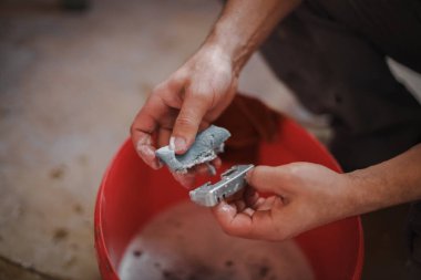 Hands of a caucasian man washes with a sponge with metal soap detail from a window frame over a red bucket of water while squatting in a room where repairs are underway, top view close-up with