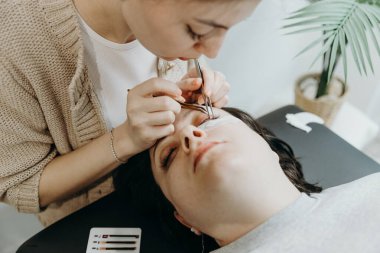 Bir kadın müşterinin sol gözündeki iki cımbızla yapay kirpikleri yapıştıran güzel, beyaz bir kız kozmetik uzmanının portresi.