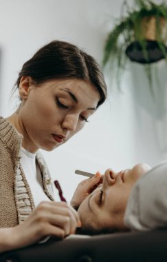 İki cımbız kullanarak dikkatle yapay kirpiklerini yapıştıran güzel beyaz bir kız kozmetik uzmanının portresi. Evdeki ofisinde kozmetoloji masasında yatan kadın bir müşteri.