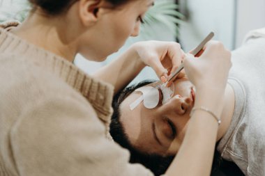 Beyaz tenli güzel bir kozmetik uzmanı kızın portresi. Yapay kirpiklerini kadın müşterisinin sol gözünde iki cımbızla yapıştırıyor. Ev ofisinin kozmetoloji masasında yatıyor.