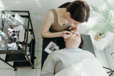 Kozmetoloji masasında yatan bir kadın müşteriye iki cımbız yardımıyla sağ gözüne yapay kirpikler yapıştıran güzel, beyaz bir kozmetik uzmanı kızın portresi.