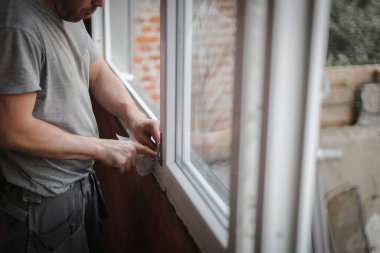 Caucasian man in gray uniform cleans his hands with dry paper napkin inside window frame opening, close up side view with selective focus. The concept of home renovation, washing window frames.
