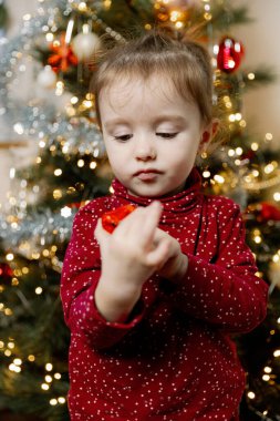 Güzel, beyaz bir kız bebek, hediyeyi ilgiyle inceliyor, bir duvar takviminin cebinden çıkarıyor, akşam vakti bulanık bir Noel ağacının yanında duruyor, oturma odasında, yakın plan.