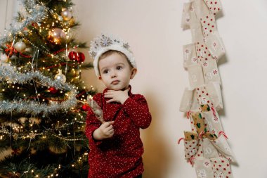 Geyik boynuzlarından oluşan boynuzları olan beyaz bir kız zarif bir Noel ağacı ile beyaz bir duvardaki takvim arasında duruyor ve cebinden bir hediye alıyor.