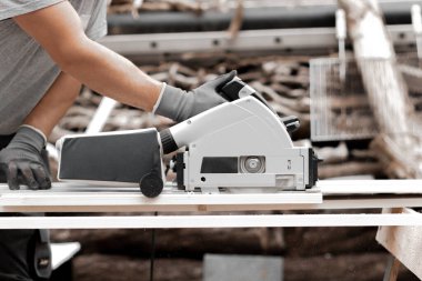 Üniformalı, gri eldivenli, tanınmayan genç bir beyaz adam bir tahtayı elektrikli testereyle kesiyor. Bir yaz günü evin arka bahçesinde dikiliyor..