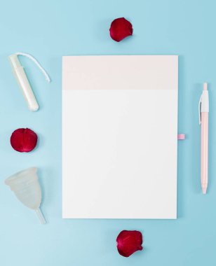 Boş defter, regl kupası, tampon, üç kırmızı gül yaprağı ve kalem mavi arka planda, düz açıda yatar. Kadın sağlığı kavramı.