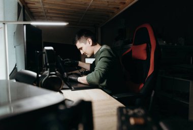 Yeşil kazaklı ve siyah eldivenli yakışıklı beyaz bir adam dikkatle bir dizüstü bilgisayara soyut koruyucu bir film yapıştırıyor, akşamları bir ofis masasında gece lambası altında oturuyor.