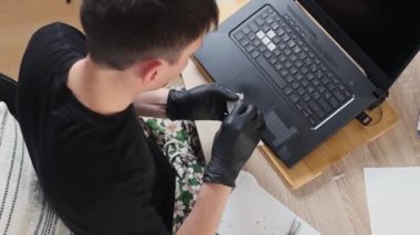 Siyah lateks eldivenli genç bir beyaz adam bir yaz günü yatak odasındaki çalışma masasında otururken dizüstü bilgisayarın klavyesine özel bir kağıt yapıştırıyor.