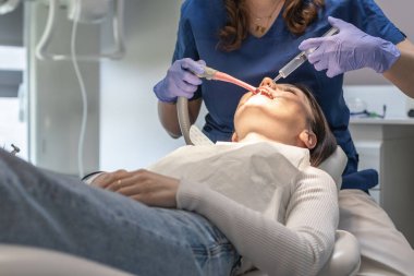 Mavi üniformalı bir dişçi ve leylak eldivenli bir dişçi dezenfektan sıvısıyla ağız boşluğunu duruluyor. Sandalyede yatan genç bir esmer hastanın şırıngasını kullanıyor.