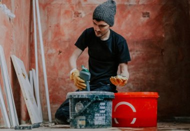 Siyah elbiseli ve sarı lastik eldivenli genç bir beyaz adam bir şişe deterjan tutuyor. Süngerin üzerine dökmeye hazırlanıyor. Restorasyonun yapıldığı odanın köşesinde dizlerinin üzerinde oturuyor.