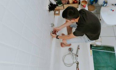 Genç bir Kafkasyalı esmer, elleriyle duvardaki musluktan yuvarlak kısımları kaldırıyor, parmaklarını kullanarak, küvetin kenarında oturuyor, üst manzara. Adım adım..