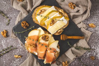 Armut, peynir, fındık, bal, kurutulmuş dilimler ve bir buğday ekmeğinin üzerinde bir dal biberiye, ortada taş tepsi, koyu beton bir masanın üzerinde, düz bir şekilde uzan. Sağlıklı beslenme