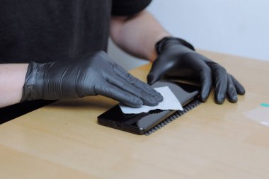 One young unrecognizable Caucasian man in black clothes and latex gloves cleans the back of a smartphone with a napkin after repairing and replacing the protective film, sitting at a wooden table in clipart