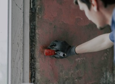 Eldivenli genç bir esmer adam kirli bir duvara fırçayla kırmızı çimento sürüyor. Küflü lekeli, yakın çekim manzaralı. Ev yenileme, inşaat işi kavramı.