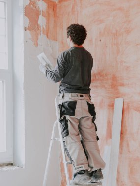 A young caucasian guy in a gray t-shirt with sleeves, work pants and curly brown hair applies fresh putty with a small spatula on the wall near the window, standing on a stepladder, close-up side view