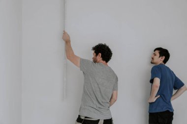Two young beautiful Caucasian plasterers in work clothes and with brown hair hold large spatulas in their hands and check the evenness of the wall after puttying in the room, close-up side view. The