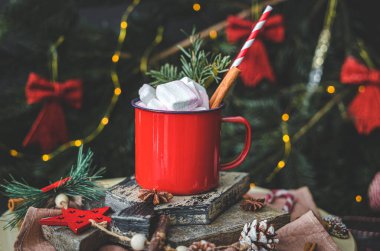 Sıcak çikolatalı bir kırmızı demir kupa, yukarıdan tarçın, pipet, kesme tahtasındaki ladin dalı ve karanlık bir şenlik arka planında süslemeleri olan noel şekeri.