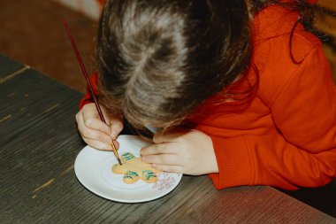 Kırmızı kazaklı, tanınmayan beyaz bir kız bir masaya oturur ve zümrüt boyayla fırınlanmış kurabiyeleri dekore etmek için eğilir. Koyu gri bir masada oturur. Adım adım.