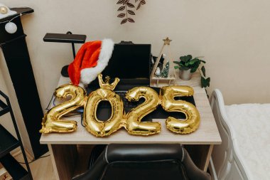 Golden inflatable numbers 2025 and santa claus hat lie right on the office table with a computer in the bedroom during the day by the black fireplace, top side view medium shot. New Year concept clipart