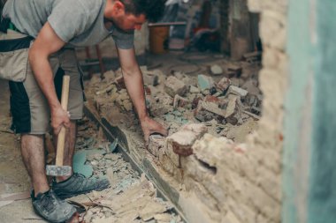 Young caucasian brunette man with a beard in work clothes breaks a brick wall with a sledgehammer in an old house,side view close-up with selective focus.Construction work concept.