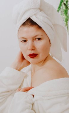 Kafasında beyaz bir havlu olan beyaz tüylü bornozlu güzel beyaz esmer bir kızın portresi beyaz bir duvarın yanında duruyor, bir omzu hafifçe açık bir şekilde yan tarafa bakıyor.