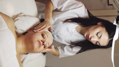 Genç bir esmer kozmetik uzmanı ellerini okşayarak yüzüne, boynuna ve omuzlarına masaj yapıyor. Güzellik masasında gözleri kapalı bir şekilde yatan bir müşteriye.