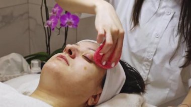 Genç bir doktor kozmetik uzmanı, bir müşterisinin sol yanağını ve alnını pembe bir süngerle temizler. Güzellik masasında gözleri kapalı bir şekilde yatar.