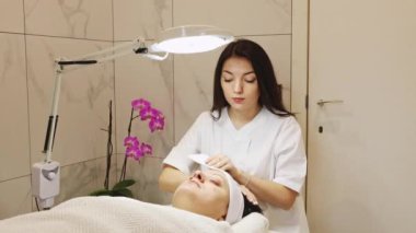 Genç ve güzel bir kozmetik uzmanı, yüz temizleme prosedürüne başlamadan önce müşterisinin kafasına beyaz bir bandaj yapıştırır. Güzellik masasında gözleri kapalı bir şekilde yatar.