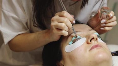 Sağ gözünün kirpiklerini silikon bir formda kaplayan, kahverengi boyayı fırçayla uygulayan beyaz bir kozmetik uzmanı. Kozmetik masasında yatan bir kadın.