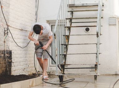 A caucasian guy in gray sweatpants and a T-shirt is leaning over washing the stairs to the terrace in the backyard of his house under the pressure of water from Karcher metal, side view, close-up. The clipart