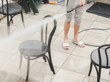 Gri eşofmanlı ve tişörtlü beyaz bir adam evinin arka bahçesinde, Karcher 'dan gelen basınçlı su jetinin altında plastikten siyah sandalyeleri yıkıyor. Yıkama konsepti