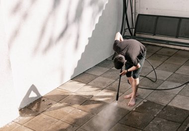 Siyah elbiseli, esmer, beyaz bir adamın portresi. Eğik bir pozisyonda yalın ayak bahçe fayanslarını baskı altında su püskürterek temizliyor. Güneş doğarken beyaz bir duvarın yanında bir kerpetenle.