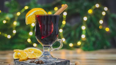 Glass of red wine mulled wine with straws, cinnamon and orange on a cutting board with a kitchen napkin on the left on a brown wooden table with a Christmas tree, red beads and a bocque garland in the clipart