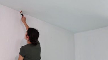 Beyaz tenli genç bir kız arkadan yan duruyor ve oturma odasında fırça ve beyaz boya ile tavanın köşelerini boyuyor, yenileme çalışmaları devam ediyor.
