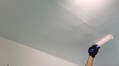 Beyaz tenli, tanınmayan bir gencin eli oturma odasındaki tavanı silindirle boyarken, yaz günü yenilenme çalışmaları devam ediyor.