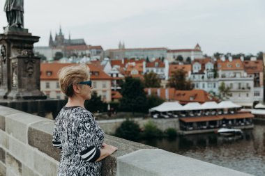 Güneş gözlüklü, sarışın, güzel, beyaz bir kadının portresi Charles Köprüsü 'nde yarım yamalak duruyor, ellerini korkuluklara dayıyor, nehrin güzelliğine bakıyor.