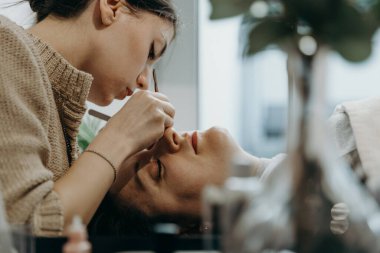 Beyaz tenli güzel bir kozmetik uzmanı kızın portresi. Yapay kirpiklerini, bir kadın müşterinin sol gözündeki iki cımbızla yapıştırıyor. Ev ofisinin kozmetoloji masasında yatıyor.
