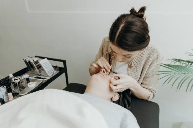 Beyaz tenli güzel kozmetik uzmanı bir kızın portresi kozmetik masasında yatan bir kadının sol göz kapağının altına cımbız yapıştırıyor.