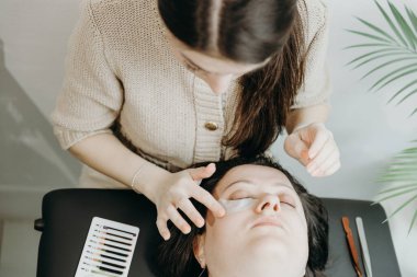 Genç ve güzel bir beyaz kozmetik uzmanı kızın portresi. Bir bahar günü ofiste kozmetoloji masasında uzanan bir kadın müşterinin sağ göz kapağının altına kağıt yapıştırıcı yapıştırıyor.