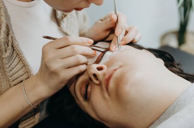 Evdeki kozmetoloji masasında yatan bir kadın müşterinin sol gözüne cımbızla yapay kirpikler yapıştıran, yarı tanınmış genç bir beyaz kız kozmetikçinin portresi.