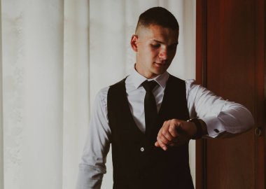 Siyah takım elbiseli mutlu beyaz damadın portresi. Kol saatine bakıyor. Yaz sabahı pencerenin yanındaki odada dikiliyor. Damat kavramı