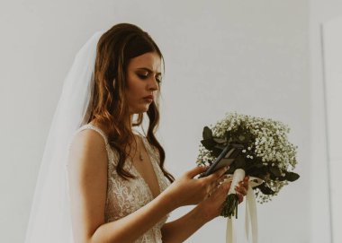 Portrait of one beautiful young Caucasian brunette bride holding a bouquet of white boutonnieres and writing a message to the grooms on a smartphone, close-up side view. clipart