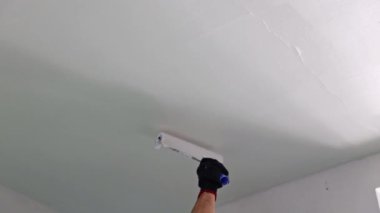 Beyaz tenli, tanınmayan bir gencin eli bir rulo boyayla resim yapıyor. Oturma odasının tavanını boyuyor. Yaz günü yenilenme devam ediyor.