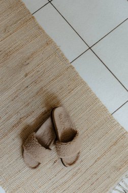 Bir çift güzel ev modasına uygun, şık ve sıcak terlikler kahve renkli doğal koyun derisi ve mantar tabanlı jüt halı üzerinde duruyor odadaki beyaz fayanslı zeminde, düz bir şekilde uzanıyor ve...