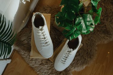 Bir çift güzel beyaz klasik spor ayakkabısı, güneşli bir günde, bir ev mağazasının kapısının yanındaki doğal yün halıda, yerde saksı çiçekli ahşap sergi kutularının üzerinde duruyor.