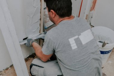 Gri üniformalı, beyaz bir genç, en alttaki kapı aralığına geniş bir mala koyarak oturuyor. Üst tarafı seçici bir odak noktasıyla yakın plan. Ev yenileme konsepti